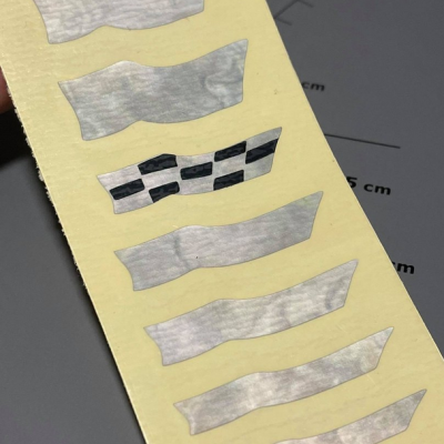 Tiras adesivas brancas e cinza amarradas a uma folha amarela, fundo cinza com régua em centímetros.