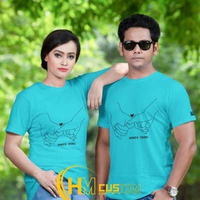 Casal com t-shirts azuis claras estampadas com mãos entrelaçadas, texto 'SINCE 1995', ao ar livre com árvores verdes.
