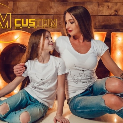Mulher e menina vestem t-shirts brancas estampadas e jeans rasgados sentadas no chão com letreiro luminoso LOVE ao fundo