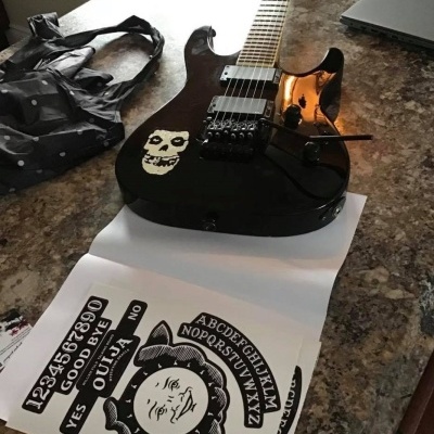 Guitarra elétrica preta com adesivo de caveira branca e folhas com desenhos de tábua Ouija numa bancada.
