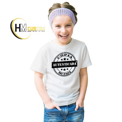 Criança vestindo camisola branca com estampa preta 'CÓPIA AUTENTICADA DO PAI' e faixa roxa na cabeça, fundo branco