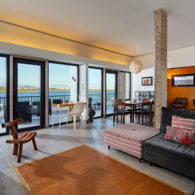 Sala de estar com sofá cinza, almofadas vermelhas e brancas, cadeira de madeira, mesa de jantar e janelas com vista para o mar