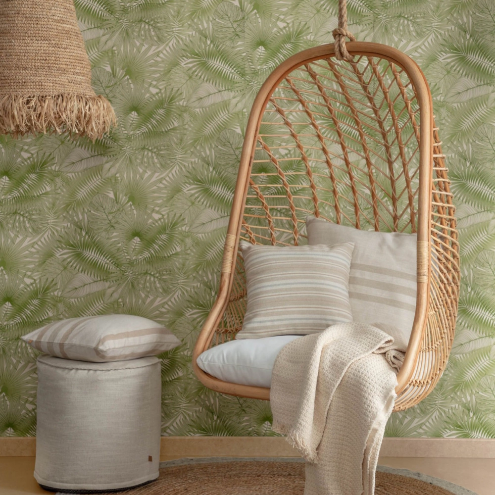 Cadeira suspensa Katerin, rattan natural Cadeira suspensa de vime com almofadas e manta junto a banco redondo cinzento e parede verde com padrão de folhas