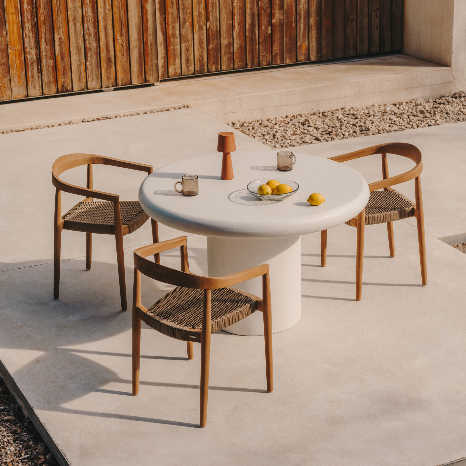 Mesa Adda, exterior, cimento, branco Mesa redonda branca com três cadeiras de madeira clara e assentos tecidos ao ar livre com limões e copos em cima.