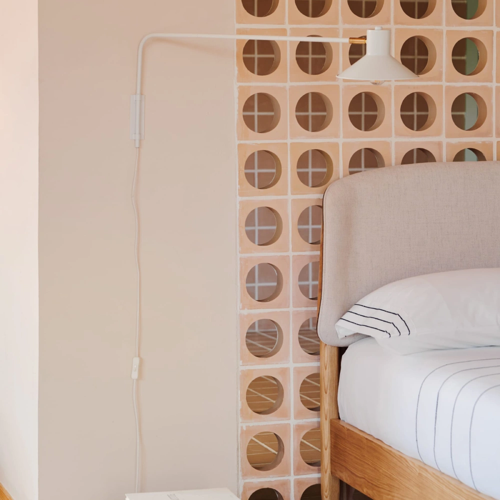 Aplique de Parede Giratório em Metal Arias Cama de madeira com cabeceira estofada bege e luminária de parede branca ao lado