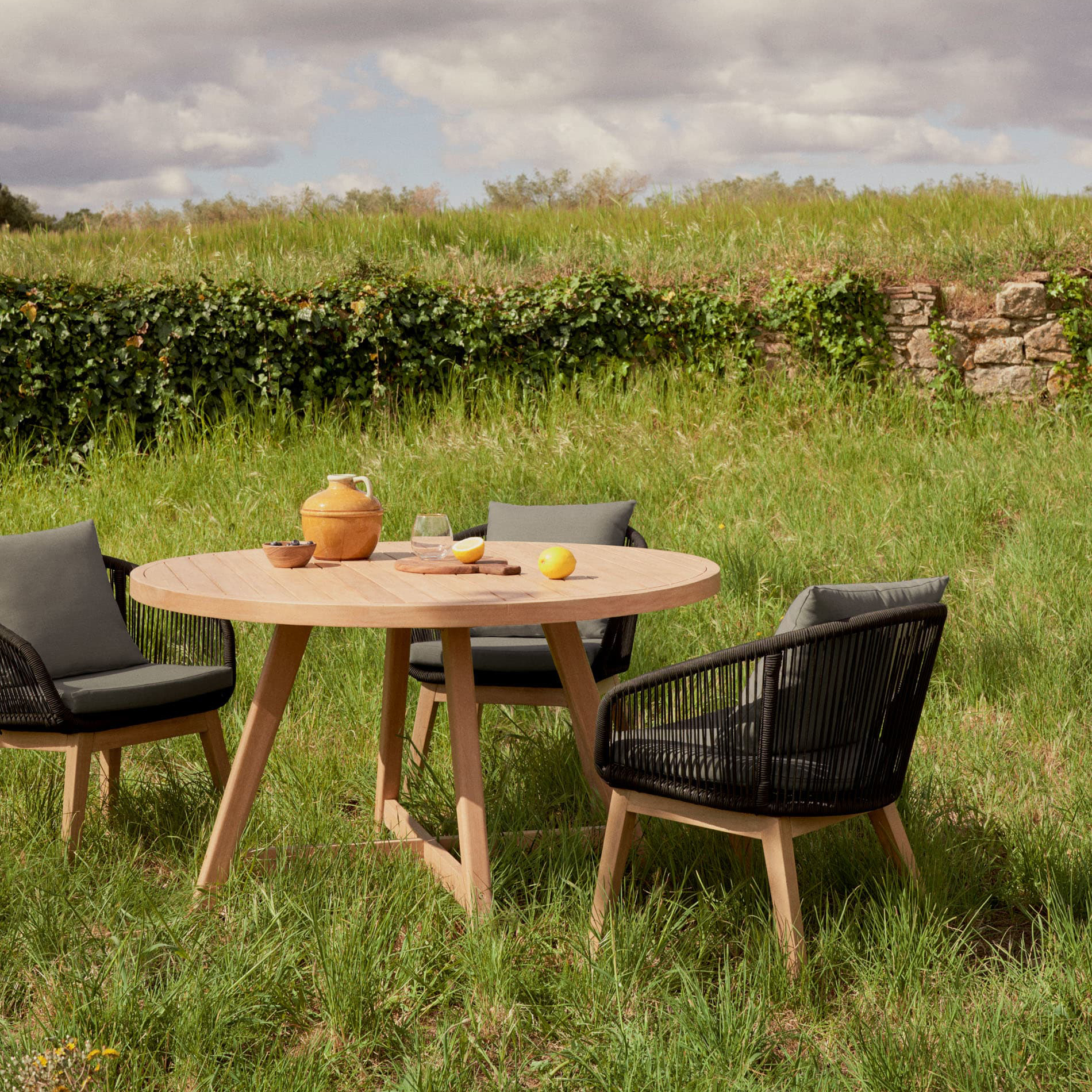 Cadeira Portale, exterior, madeira acácia Mesa redonda de madeira clara com três cadeiras em madeira e corda preta num relvado ao ar livre