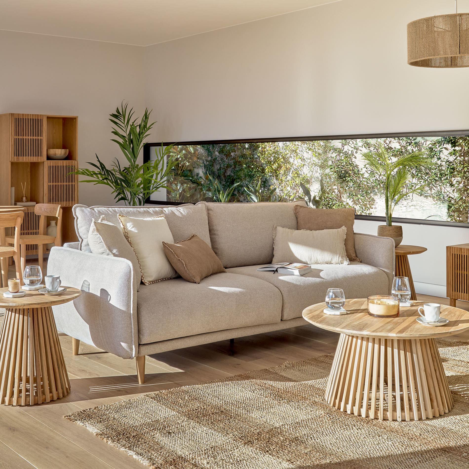 Sofá Karla, 3 lugares, estofado, bege Sofá cinzento claro com almofadas e mesas de centro de madeira num ambiente luminoso e decorado com plantas.