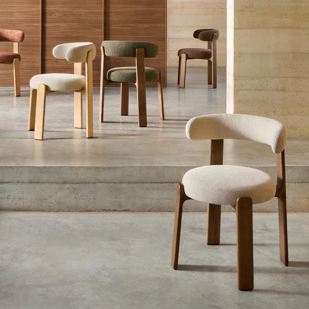 Cadeira em madeira de freixo estofada chenille Grani Sete cadeiras estofadas de diferentes cores com pernas de madeira num espaço interior com chão e paredes de cimento.