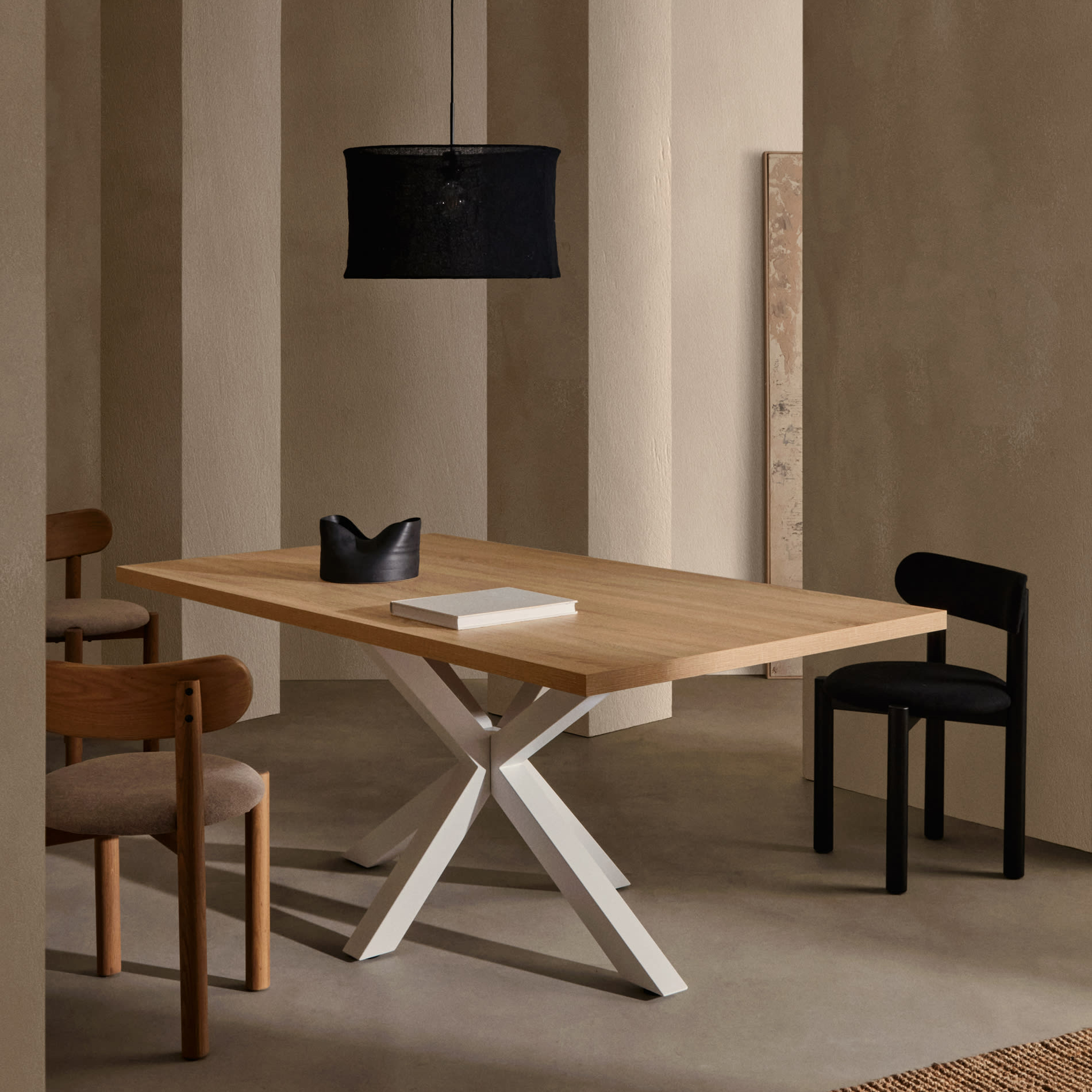 Mesa de jantar Arge, melamina/metal, branco Mesa de madeira clara com base branca em cruz, cadeiras variadas em madeira com estofos bege e preto, lâmpada preta suspensa, ambiente com paredes claras e piso cinzento.