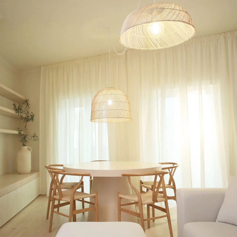 Cadeira Wishbone com braços em madeira olmo e corda natural Mesa de jantar redonda branca com cadeiras de madeira e luminárias pendentes em sala com cortinas brancas e sofá