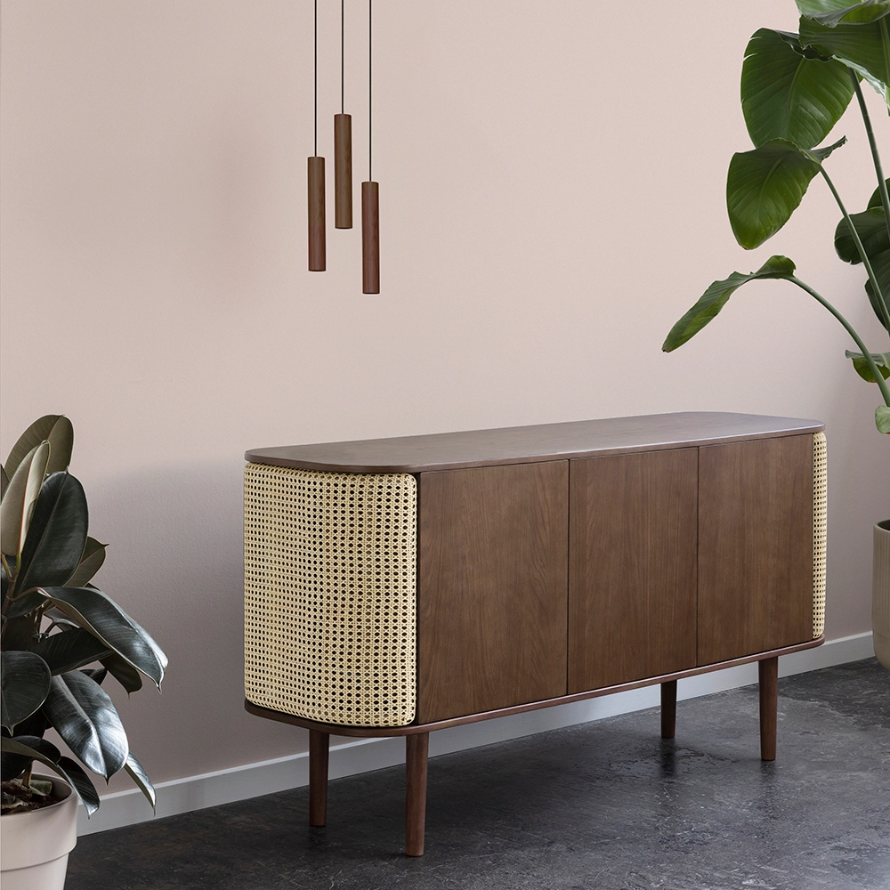 Candeeiro de Teto Chimes Cluster em Madeira de Carvalho Aparador de madeira com laterais em palha e pés cilíndricos, decorado com plantas e candeeiros suspensos.