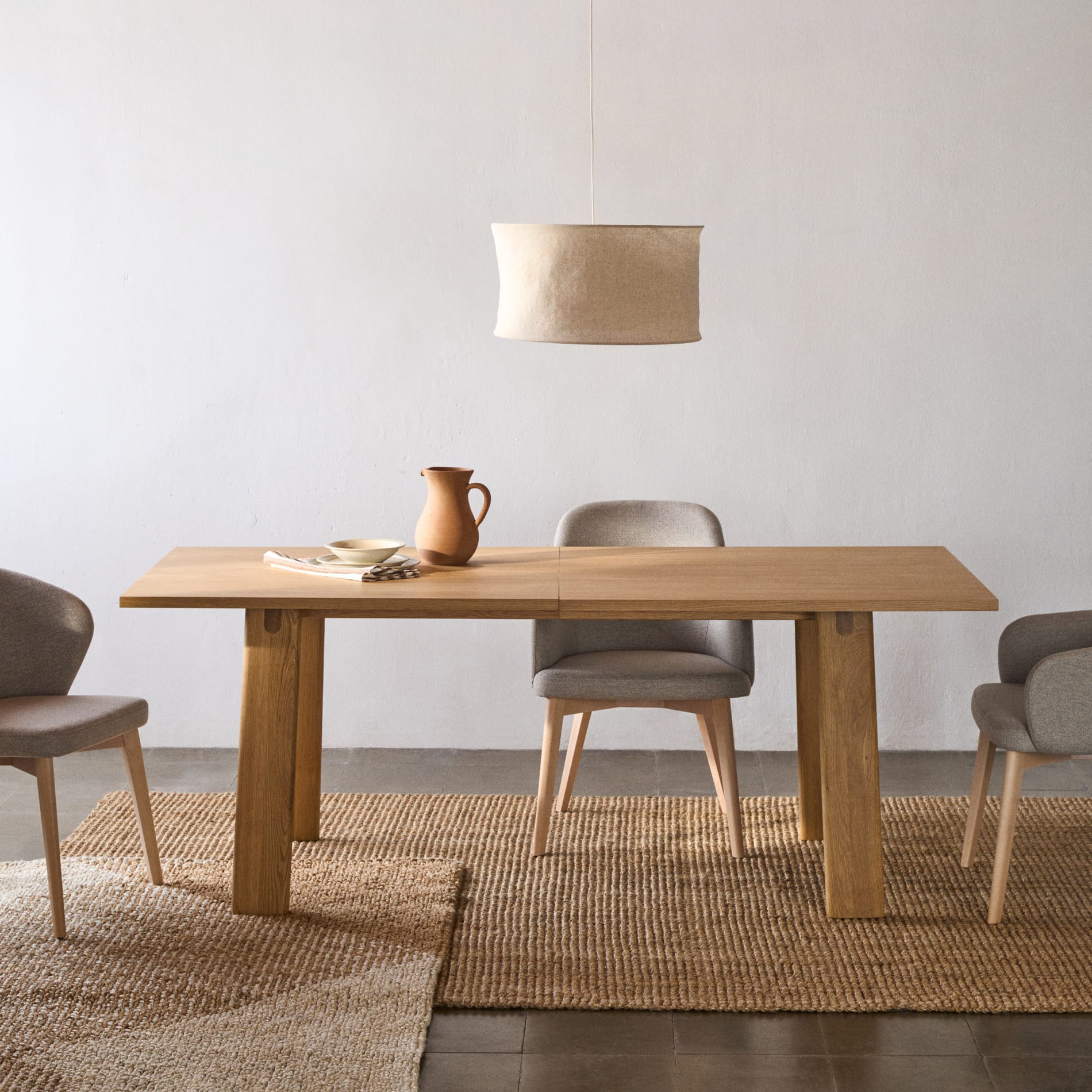 Mesa extensível Allen, madeira carvalho, 200(250)x95 Mesa de madeira clara com cadeiras estofadas cinzentas e candeeiro suspenso de tecido bege num ambiente minimalista
