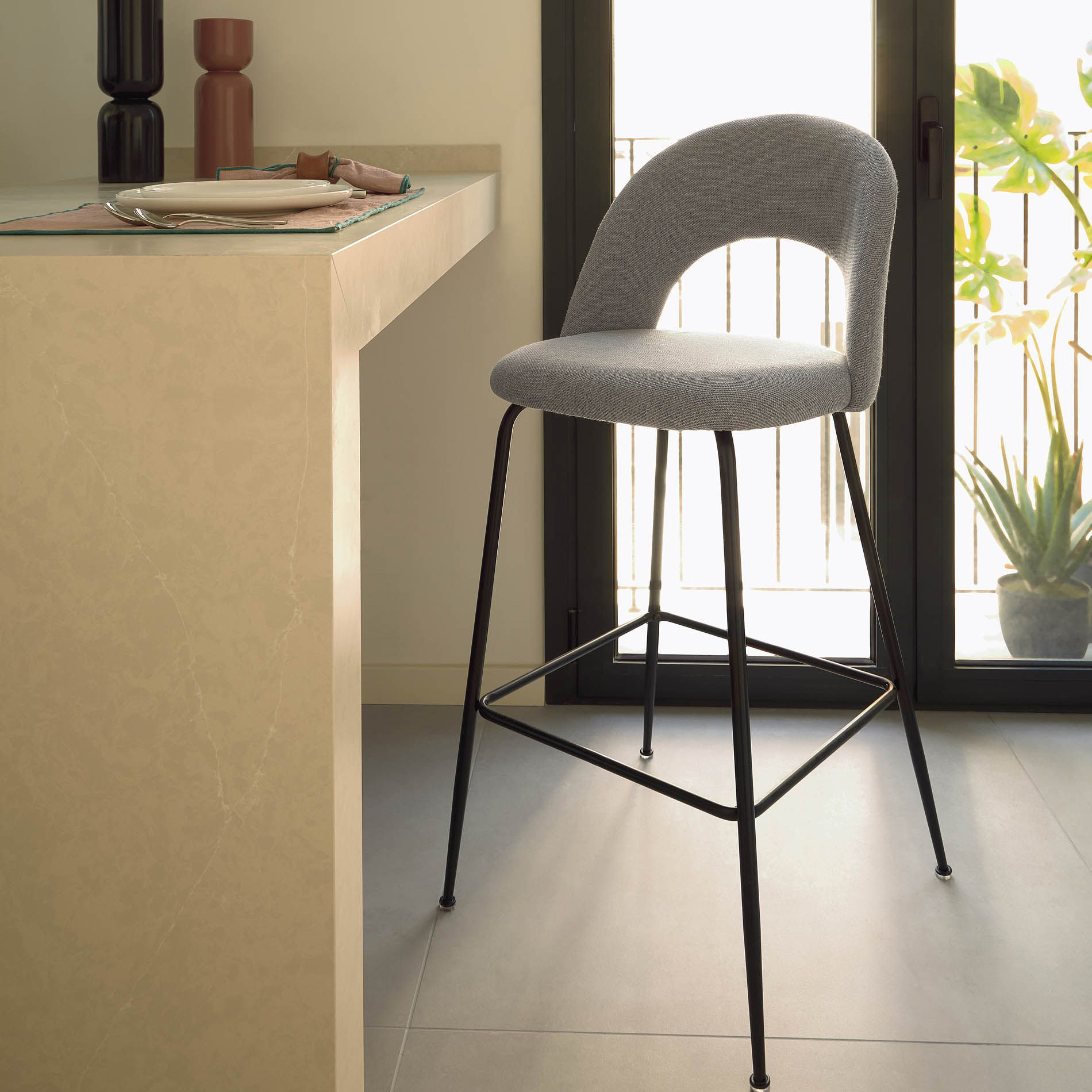 Cadeira alta Mahlia, estofada, 63cm Cadeira alta cinzenta com pernas pretas ao lado de bancada clara e portas de vidro