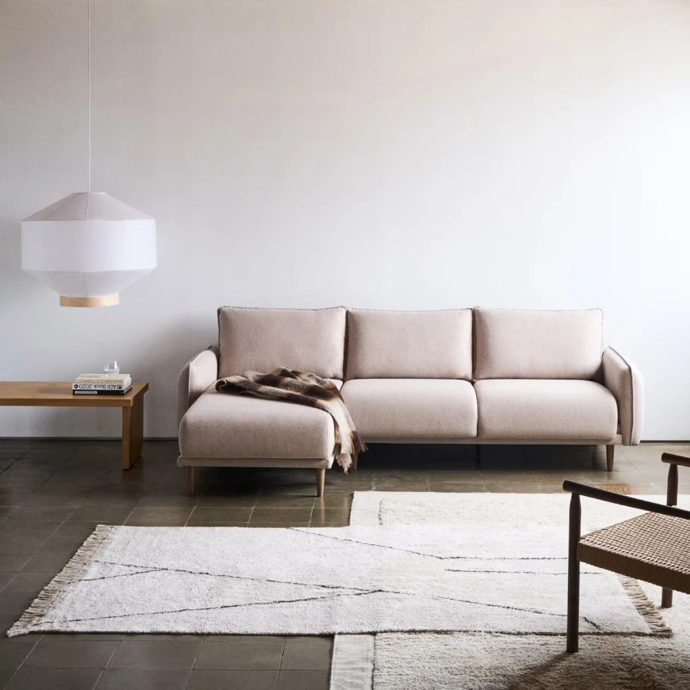 Sofá estofado bege com chaise longue direita/esquerda Karla Sofá modular bege claro com manta, mesa de madeira, candeeiro branco e tapete geométrico branco