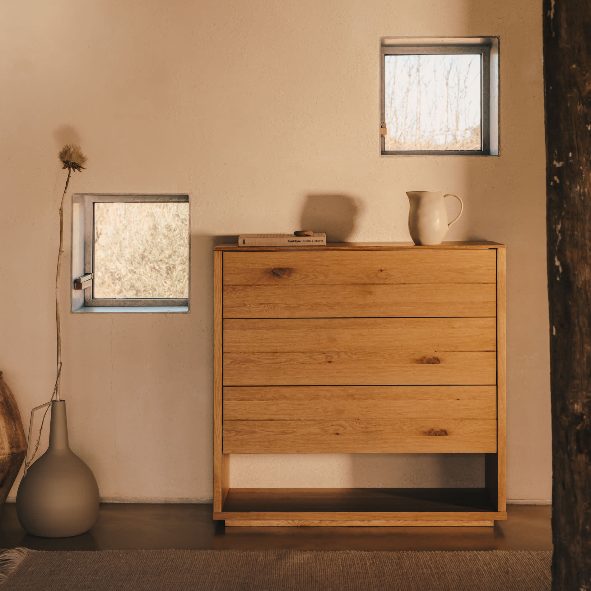 Cómoda Alguam, madeira carvalho, 100x96 Cómoda de madeira clara com jarro e livro em cima, vaso cinzento com planta ao lado, parede clara com duas janelas pequenas