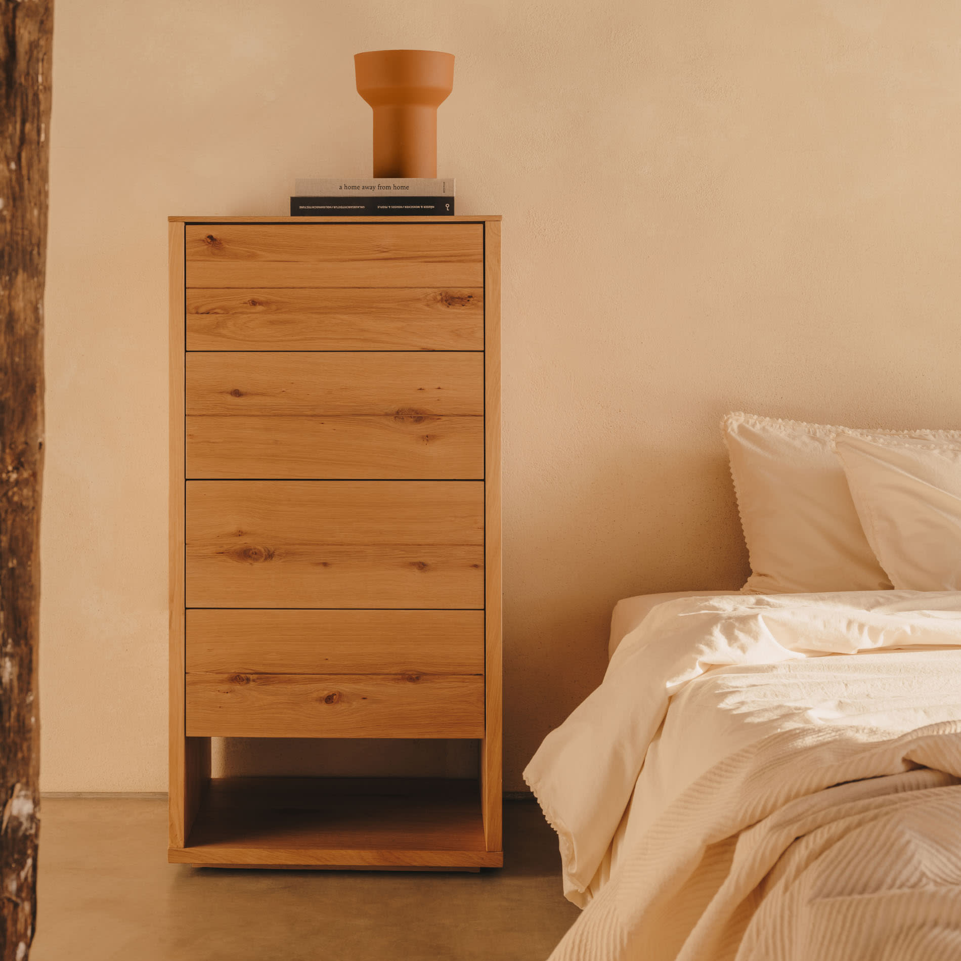 Cómoda Alguam, madeira carvalho, 60x120 Cômoda alta em madeira clara juntamente com vaso terracota e livros ao lado de cama com roupa creme.