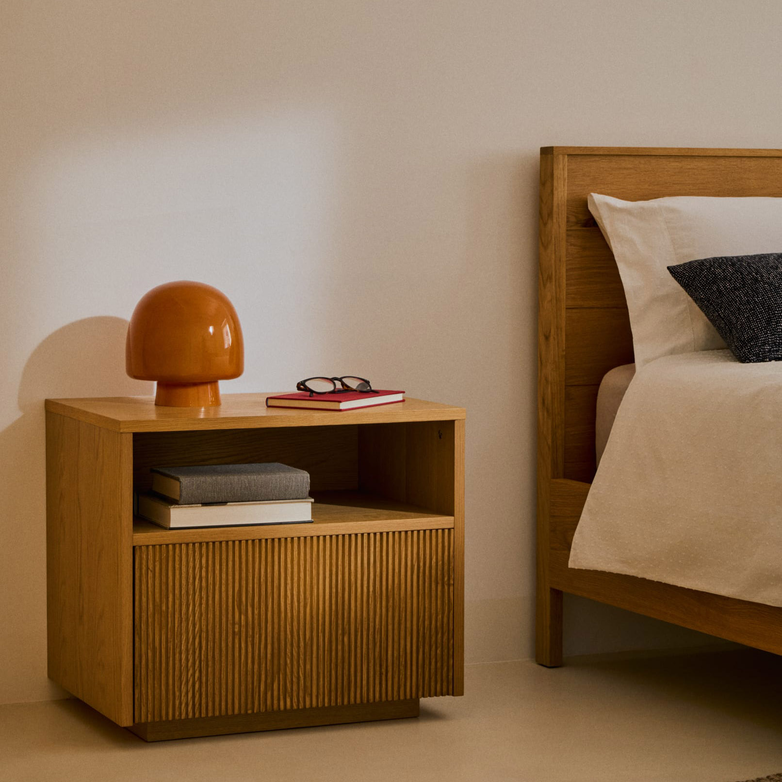 Mesa de cabeceira Helvi, madeira carvalho, 60x52 Mesa de cabeceira de madeira com abajur laranja, livros e óculos ao lado de cama de madeira com roupa branca.
