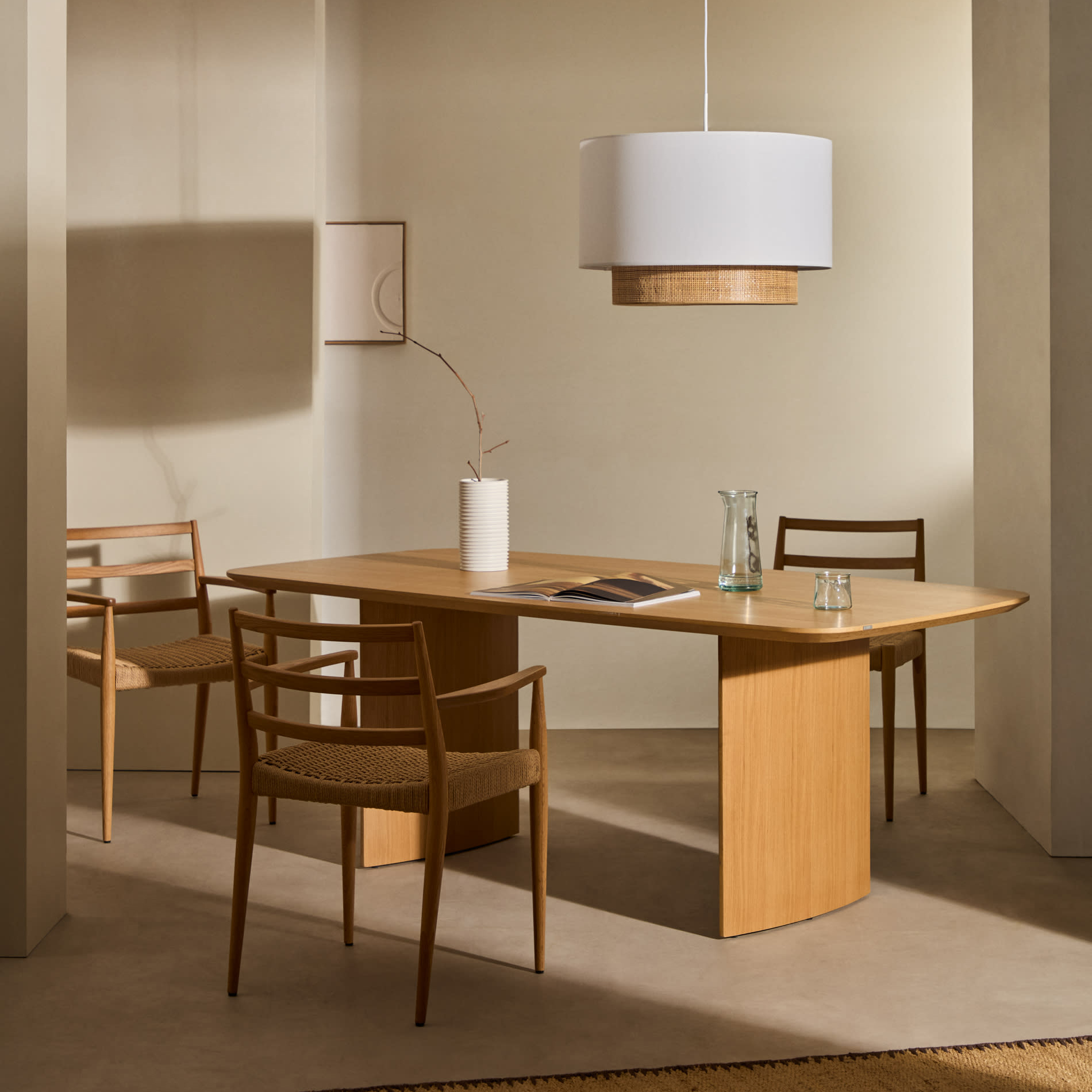 Mesa de jantar Lutto, madeira carvalho Mesa de madeira clara com quatro cadeiras castanhas, vaso branco e jarra de vidro, sob lâmpada pendente branca