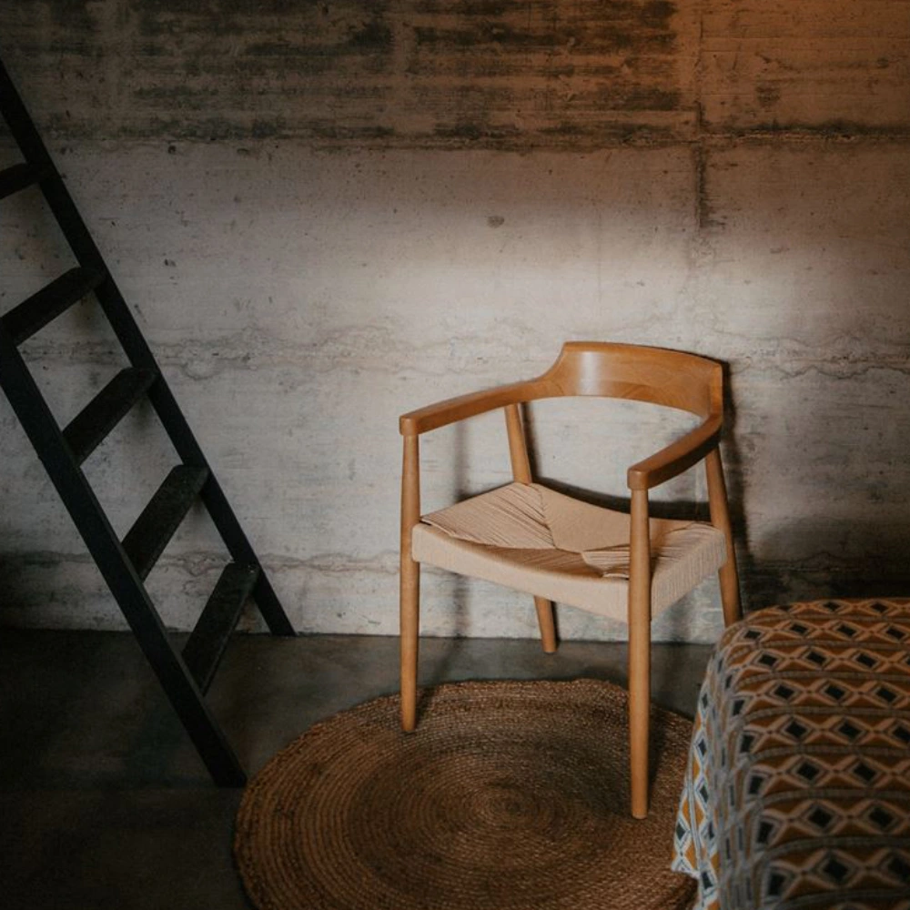 Cadeira com braços em madeira e corda bege Vagram Cadeira de madeira clara com assento trançado de corda bege em ambiente com escada preta, tapete redondo e cama com colcha geométrica.