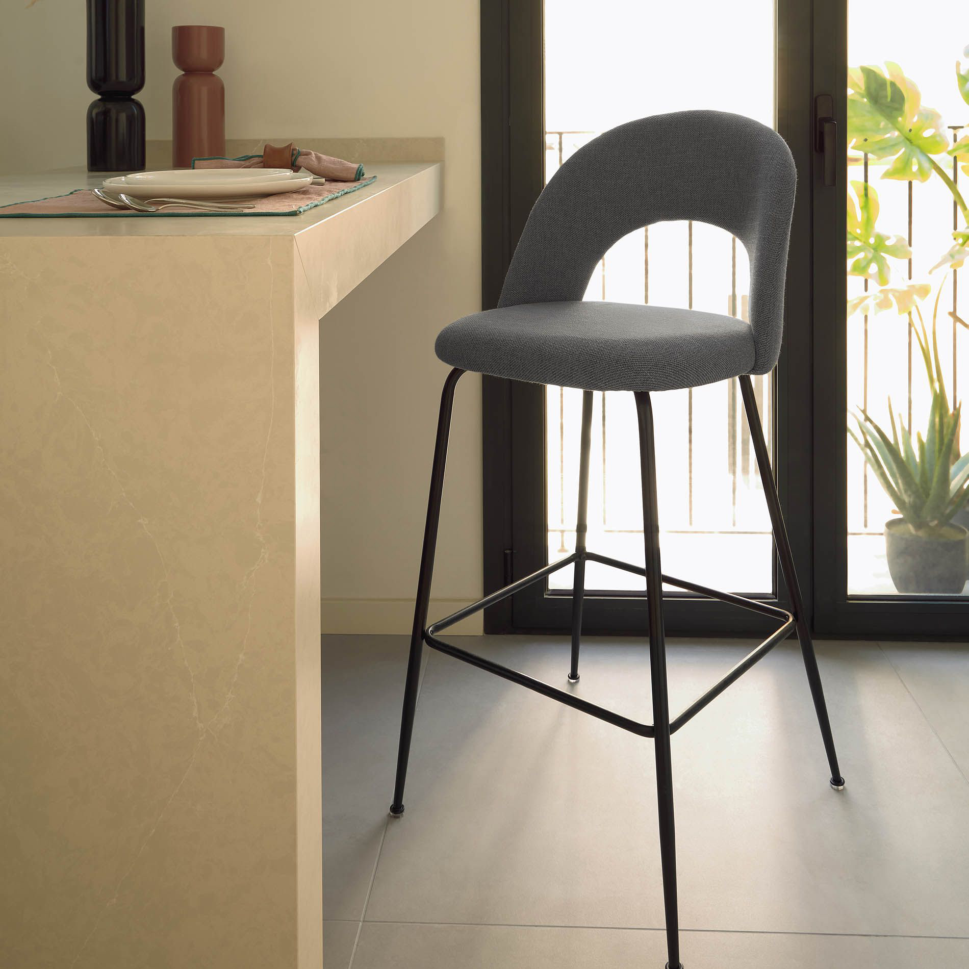 Cadeira alta Mahlia, estofada, 63cm Banqueta alta com assento cinzento e estrutura preta junto a balcão bege e porta de vidro