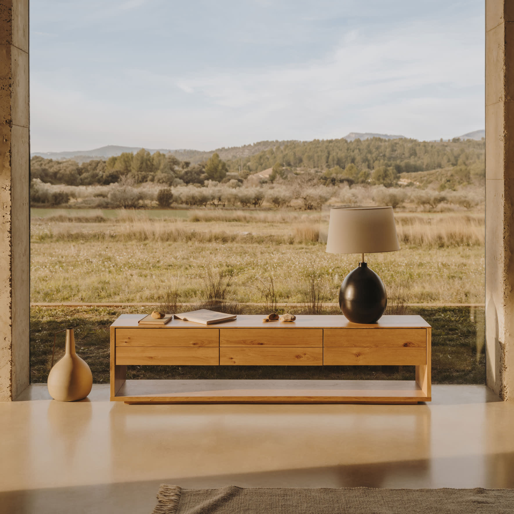 Móvel TV Alguam, madeira carvalho, 200x50 Aparador de madeira clara com candeeiro preto e vaso bege junto a janela com vista campestre