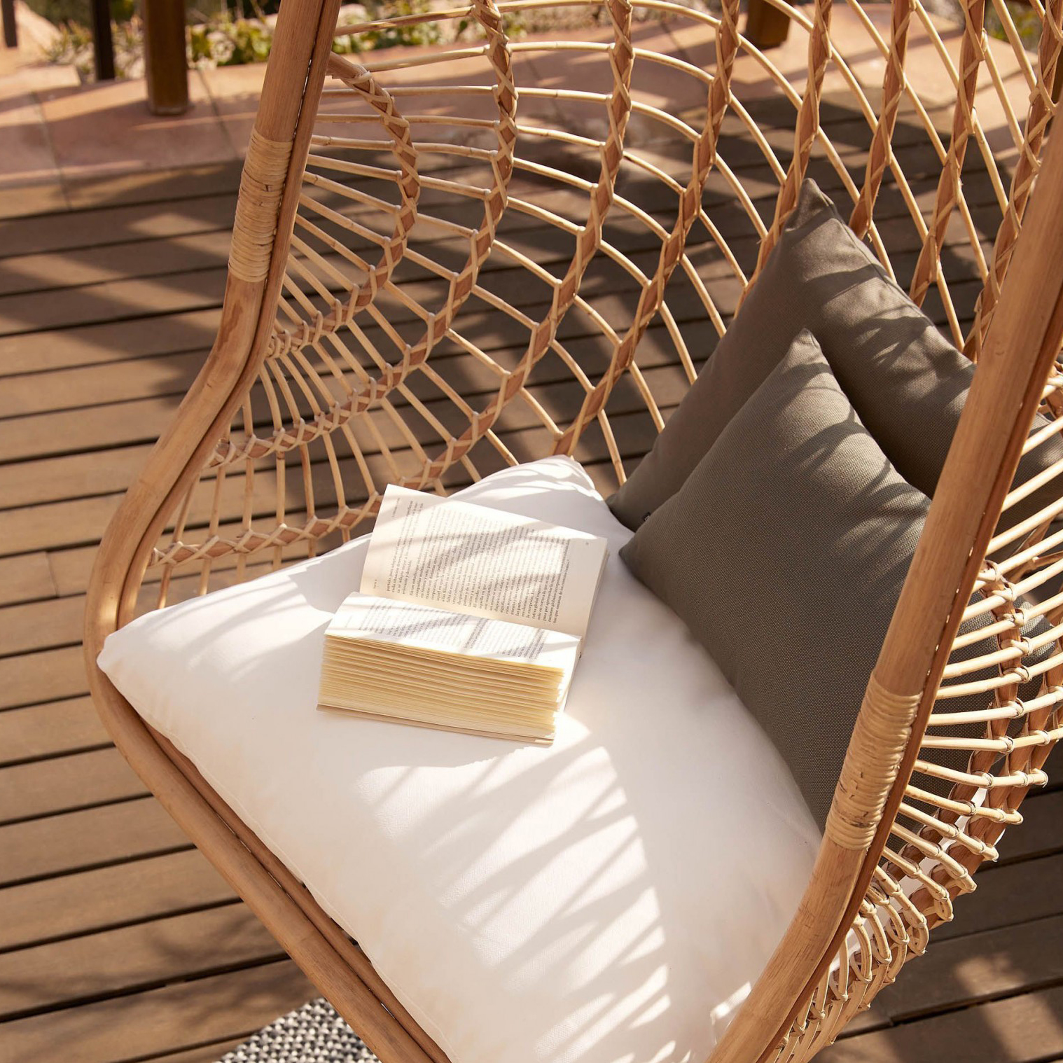 Cadeira suspensa Katerin, rattan natural Cadeira suspensa de madeira com almofadas e livro aberto num deck de madeira