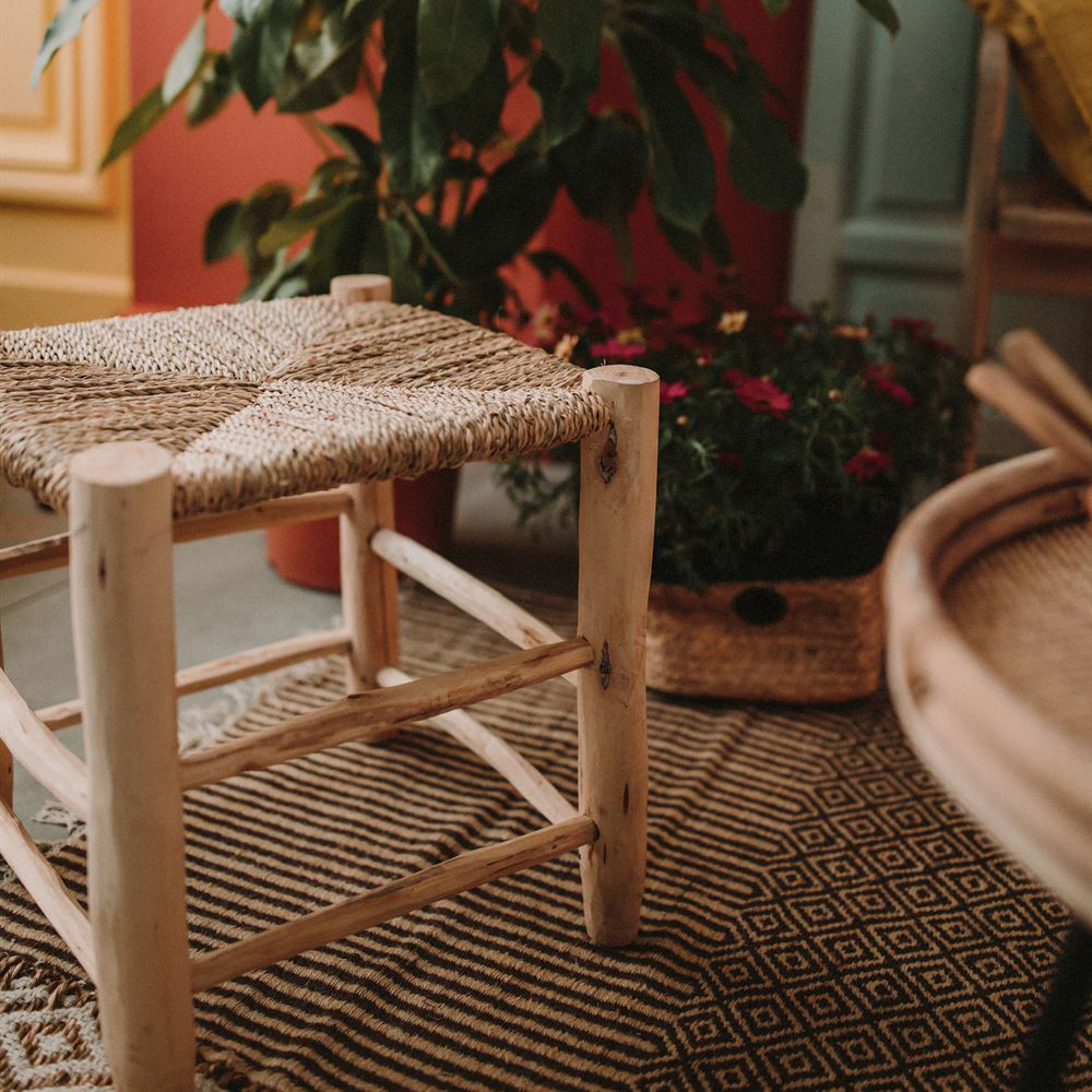 Banco de apoio Kandi, madeira/folha de palmeira Banco de madeira clara com assento tecido em corda natural, sobre tapete geométrico.