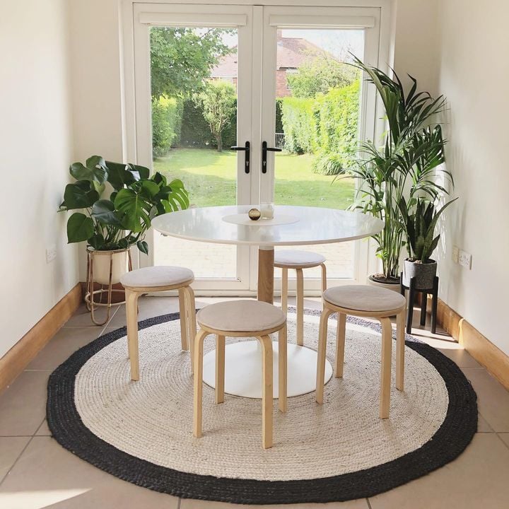 Mesa de jantar Brick, madeira de freixo/lacado, Ø110x75 Conjunto de mesa e bancos em madeira clara sobre tapete redondo branco e preto em divisão com porta de vidro e vista para jardim