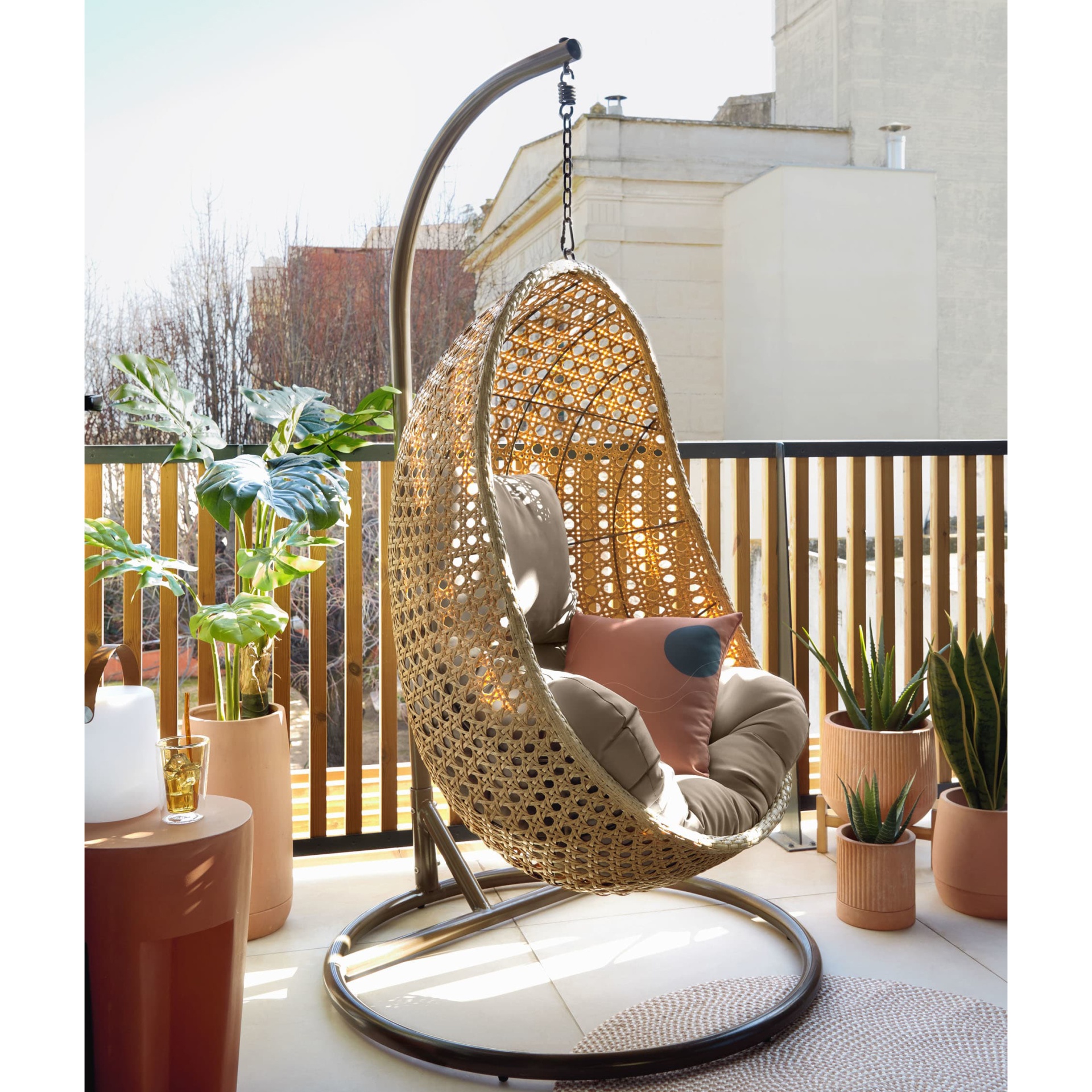 Cadeira suspensa Siran, rattan sintético, bege Cadeira suspensa em rattan sintético bege numa varanda com plantas e estrutura metálica