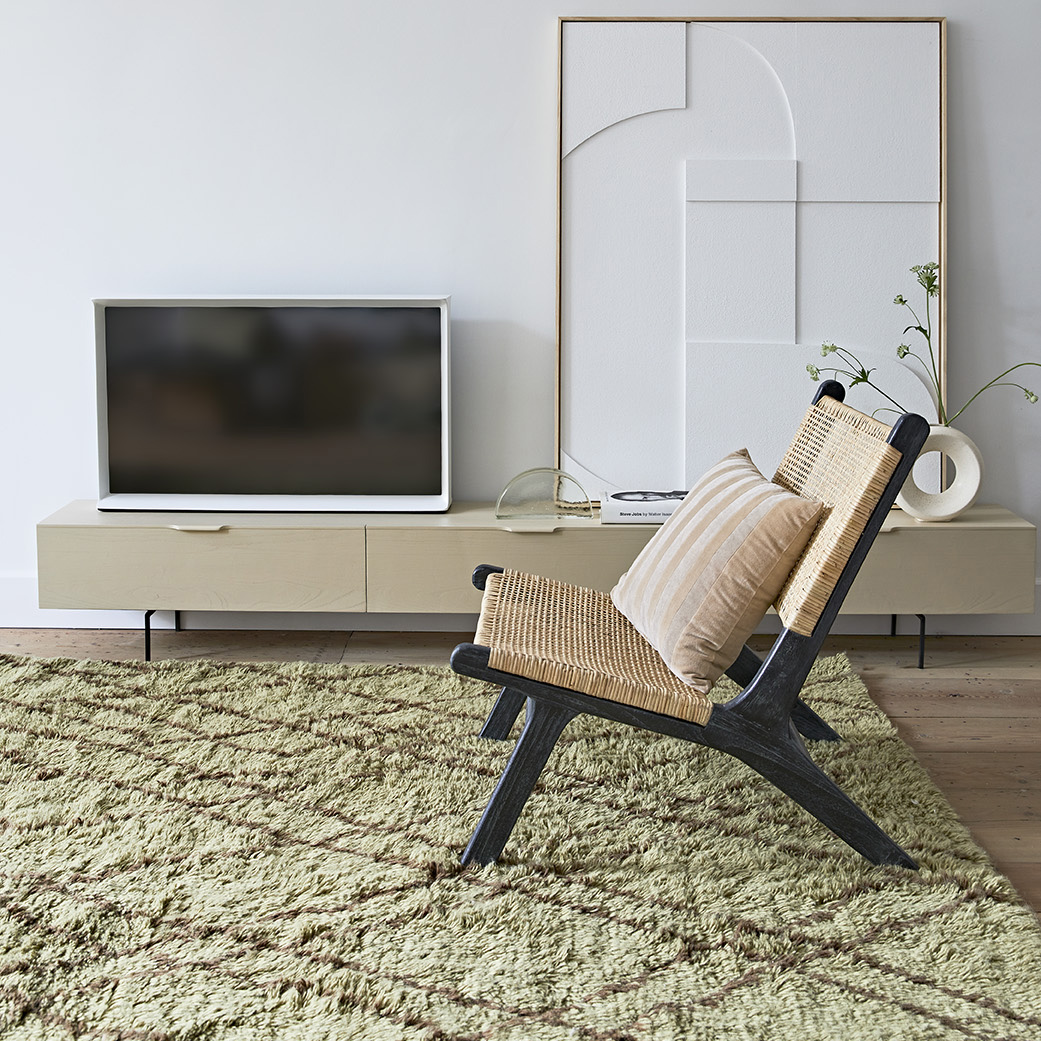 Móvel TV Wood Grain, madeira sungkai, bege, 250x36 Poltrona de madeira preta com assento de corda e almofada bege numa sala com tapete verde e móvel suspenso creme.