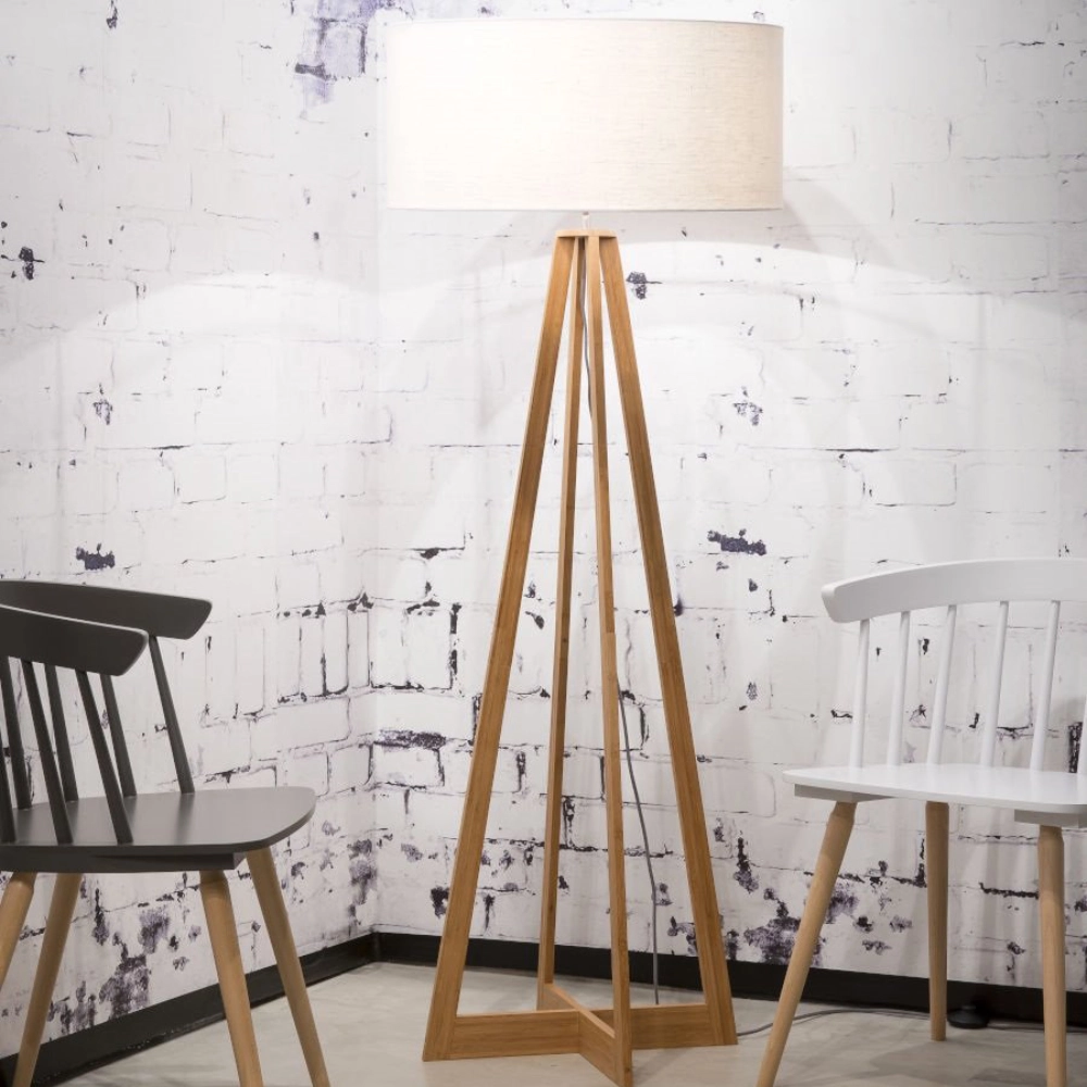 Candeeiro de Pé em Bambu e Linho Everest Lâmpada de pé de madeira com abajur branco entre duas cadeiras de madeira, preta e branca, junto a parede de tijolo branco