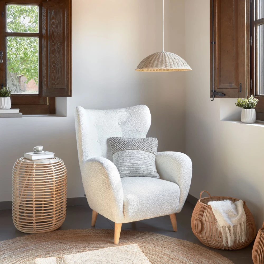 Poltrona estofada bouclé branco Pati Poltrona branca de tecido boucle com almofada cinza, mesa lateral de vime com livros e chávena, candeeiro de rattan, cesta de vime com manta e janelas de madeira com plantas.