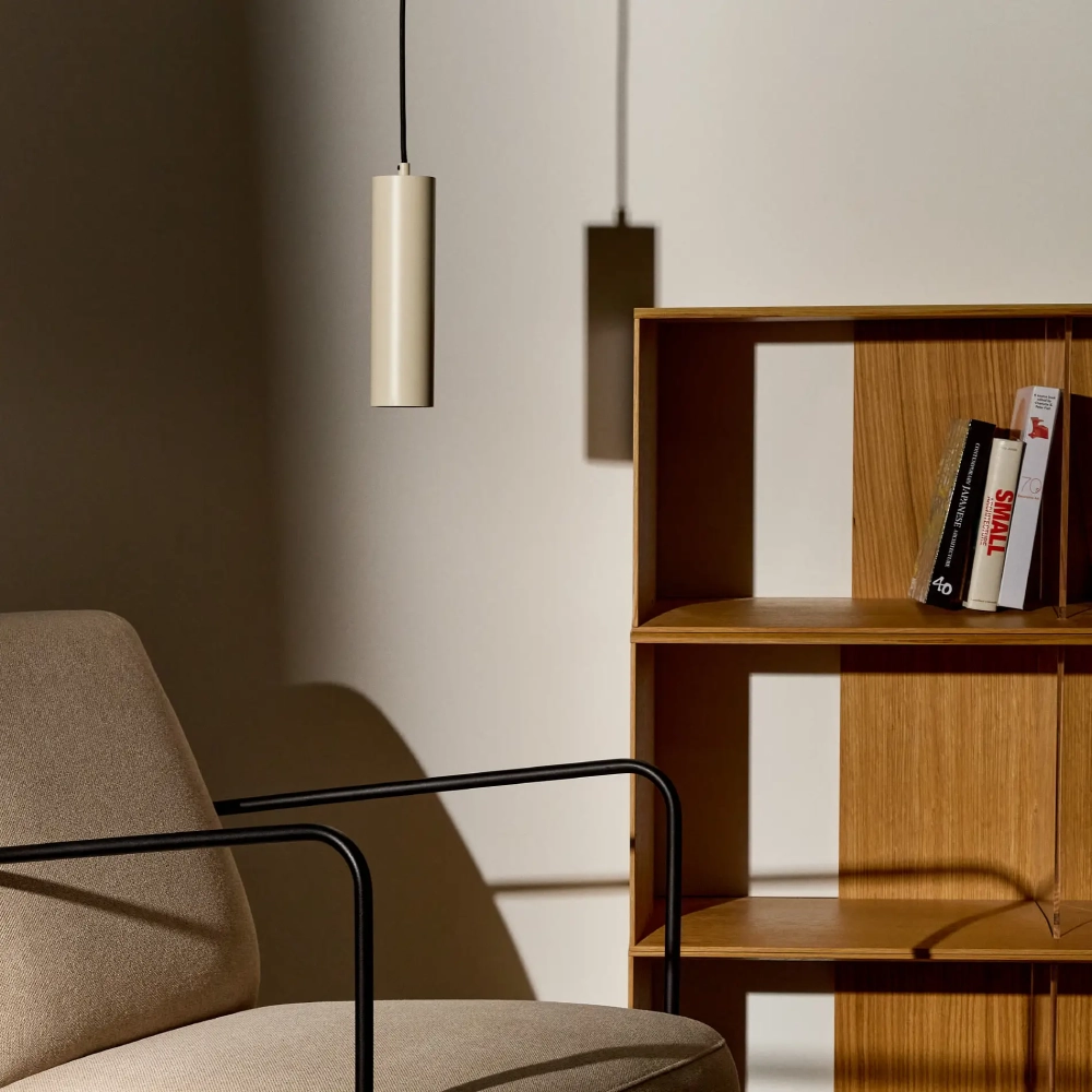 Candeeiro de teto em alumínio Adone Ø7x25 Cadeira bege estofada com braços de metal preto junto a estante de madeira com livros e luminária pendente cinza clara