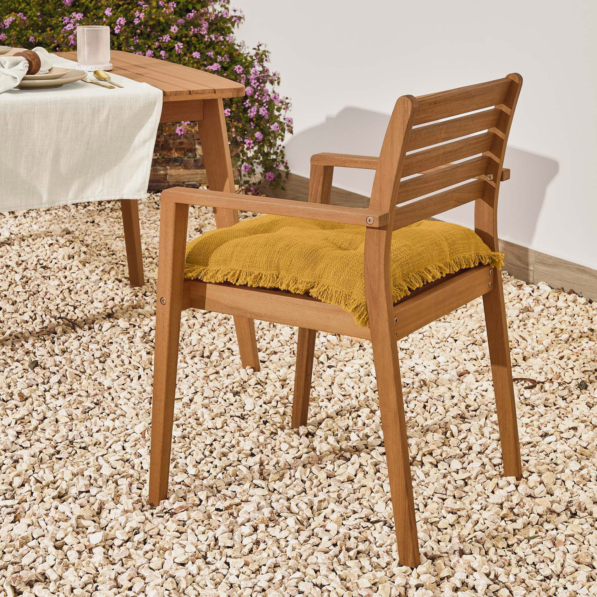 Cadeira Hanz, exterior, madeira acácia Cadeira de madeira com almofada amarela em piso de seixos ao lado de mesa com toalha branca