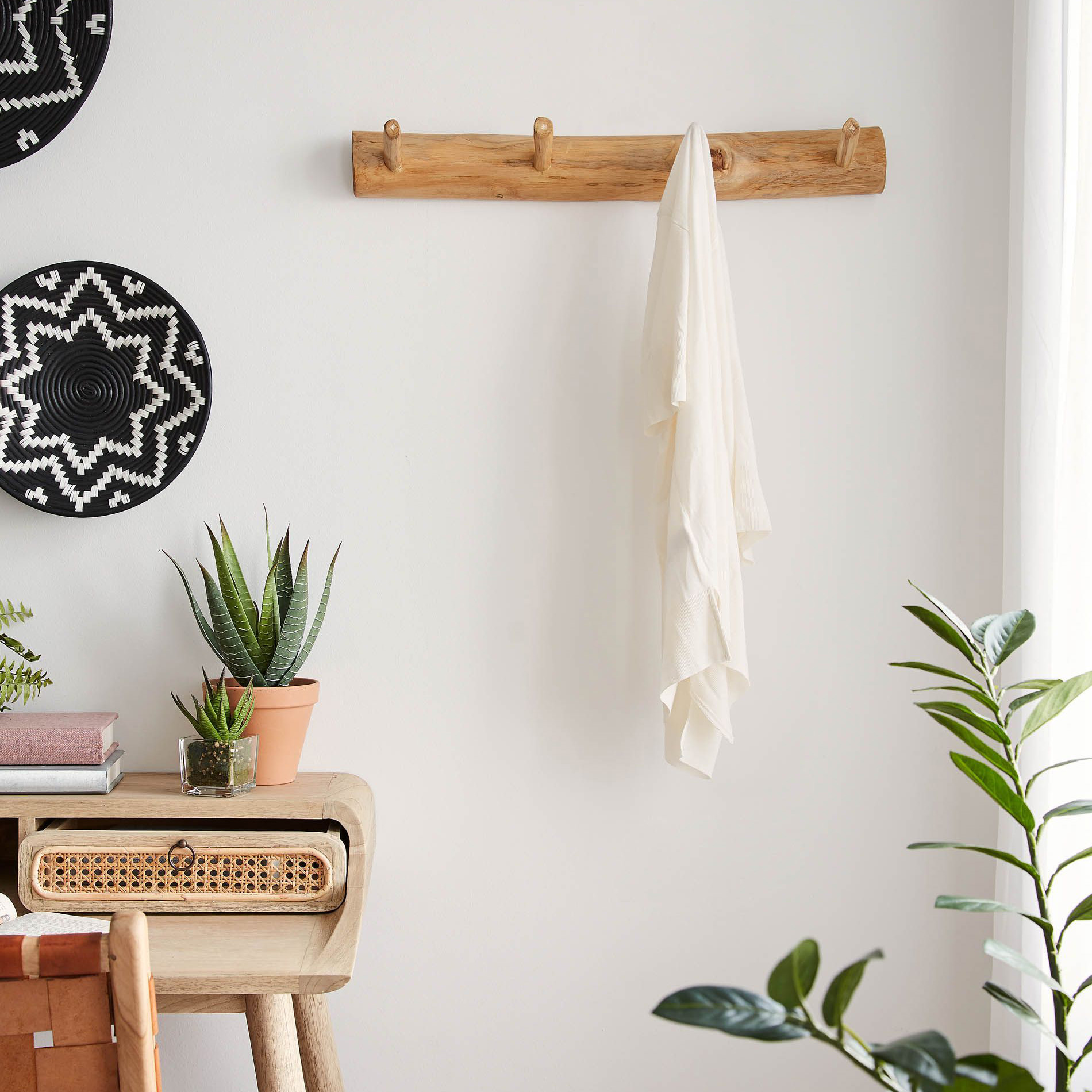 Cabide de parede Gaill, madeira teca, 63x7 Cabideira de madeira com tecido pendurado numa parede branca, perto de mesa com plantas e decoração