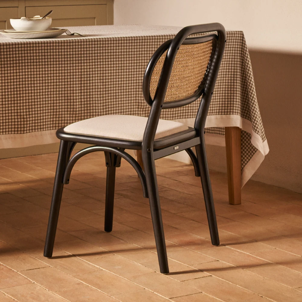 Cadeira com assento estofado bege em carvalho Doria Cadeira preta com assento branco e encosto em rattan junto a mesa com toalha xadrez