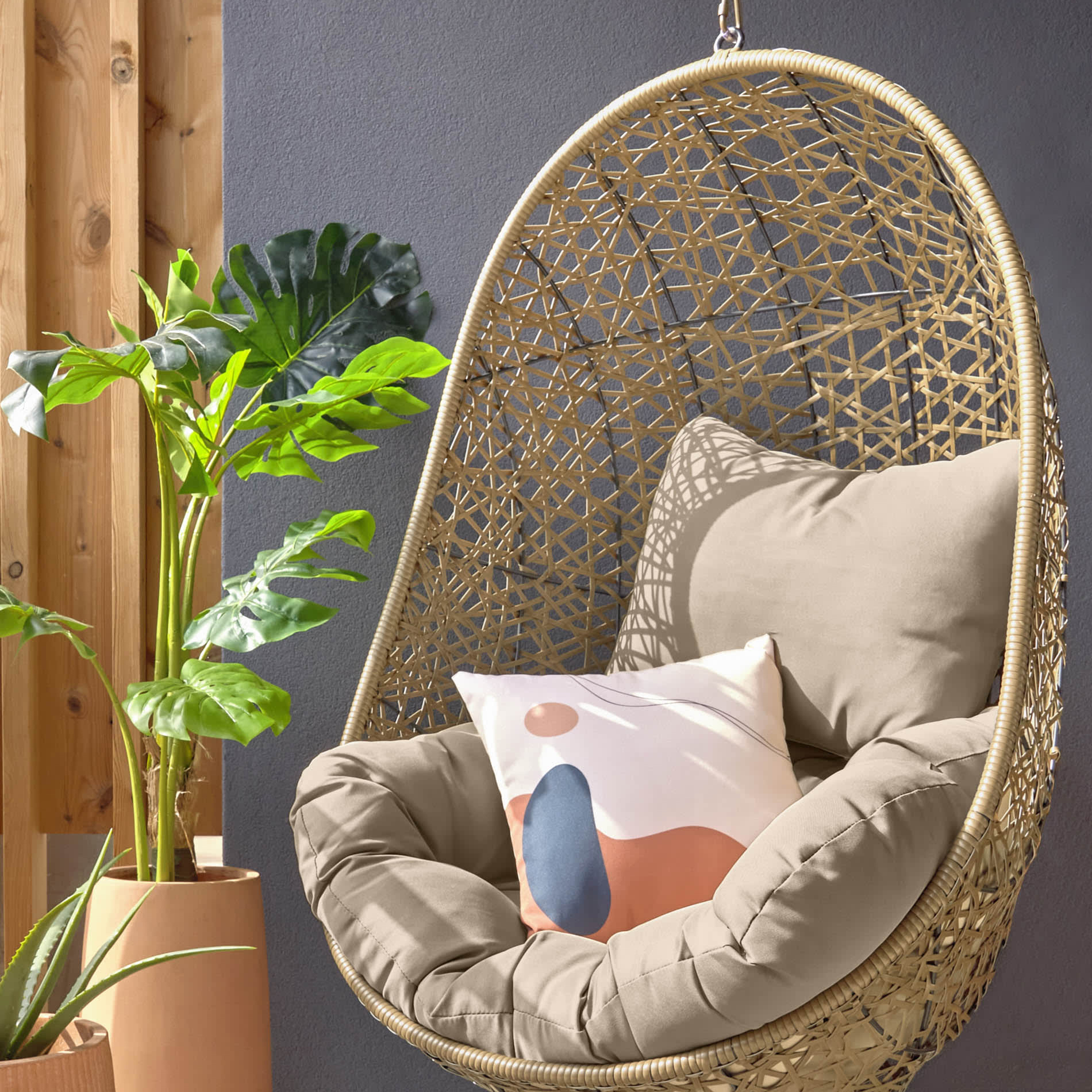 Cadeira suspensa Flora, rattan sintético, castanho Cadeira suspensa de junco com almofadas junto a plantas e parede cinza