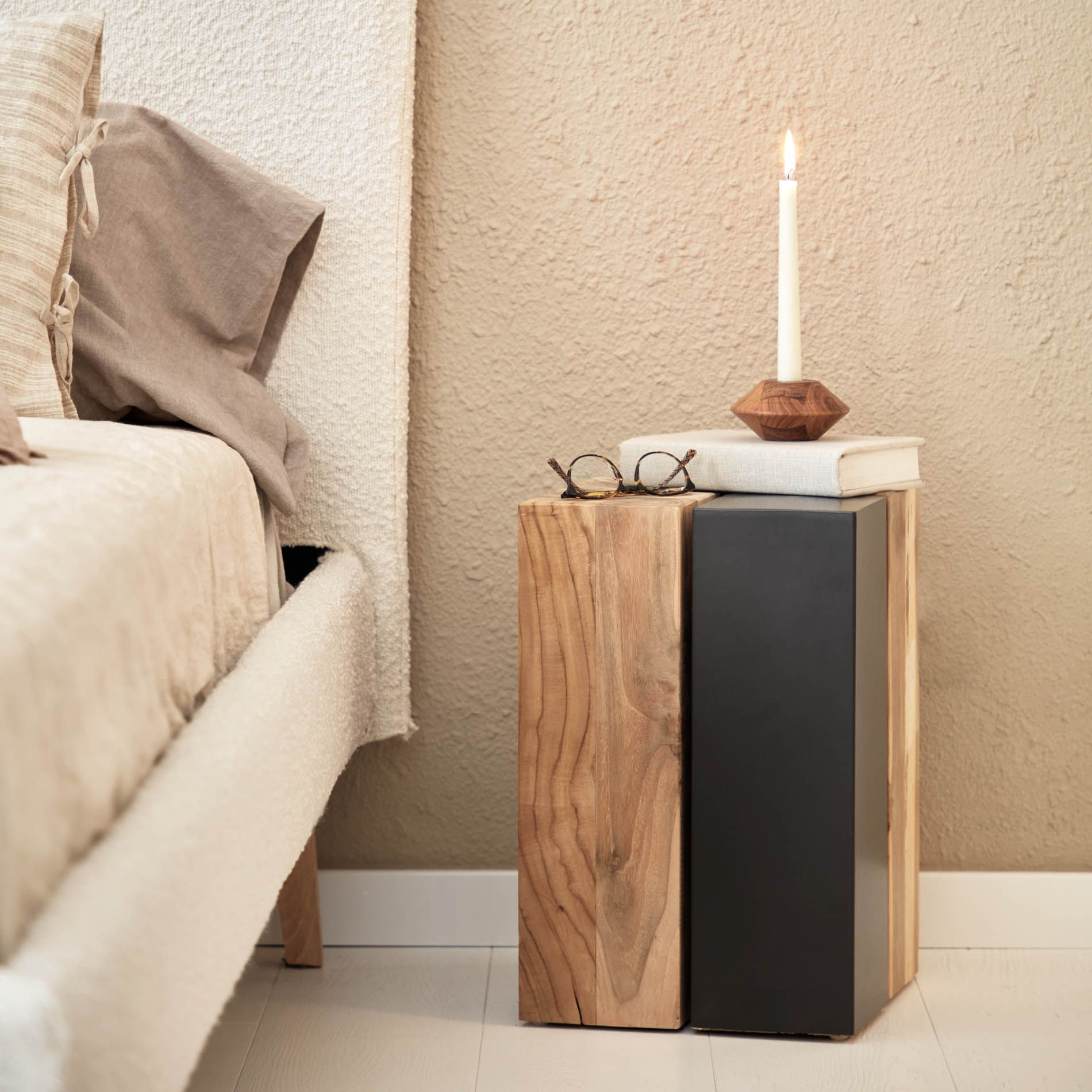 Mesa lateral Mango, madeira teca Mesa de cabeceira com bloco de madeira e bloco preto, ao lado de cama bege, com óculos, livro e vela acesa em cima.