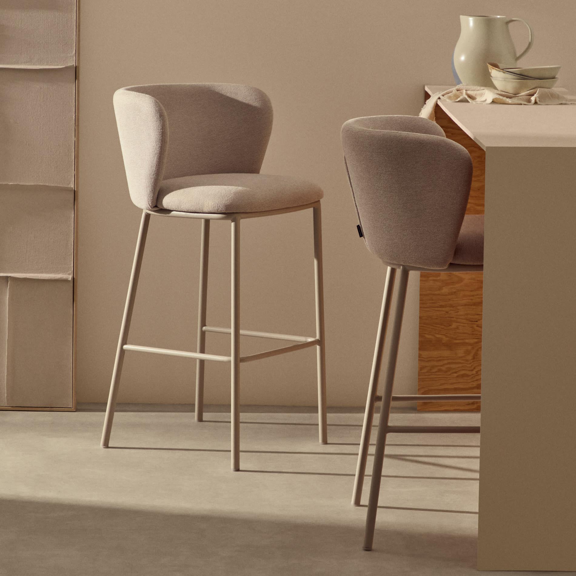 Cadeira alta Cilia, estofada, chenille, bege, 65cm Dois bancos altos estofados em tecido cinza claro com estrutura metálica bege junto a balcão de cozinha bege