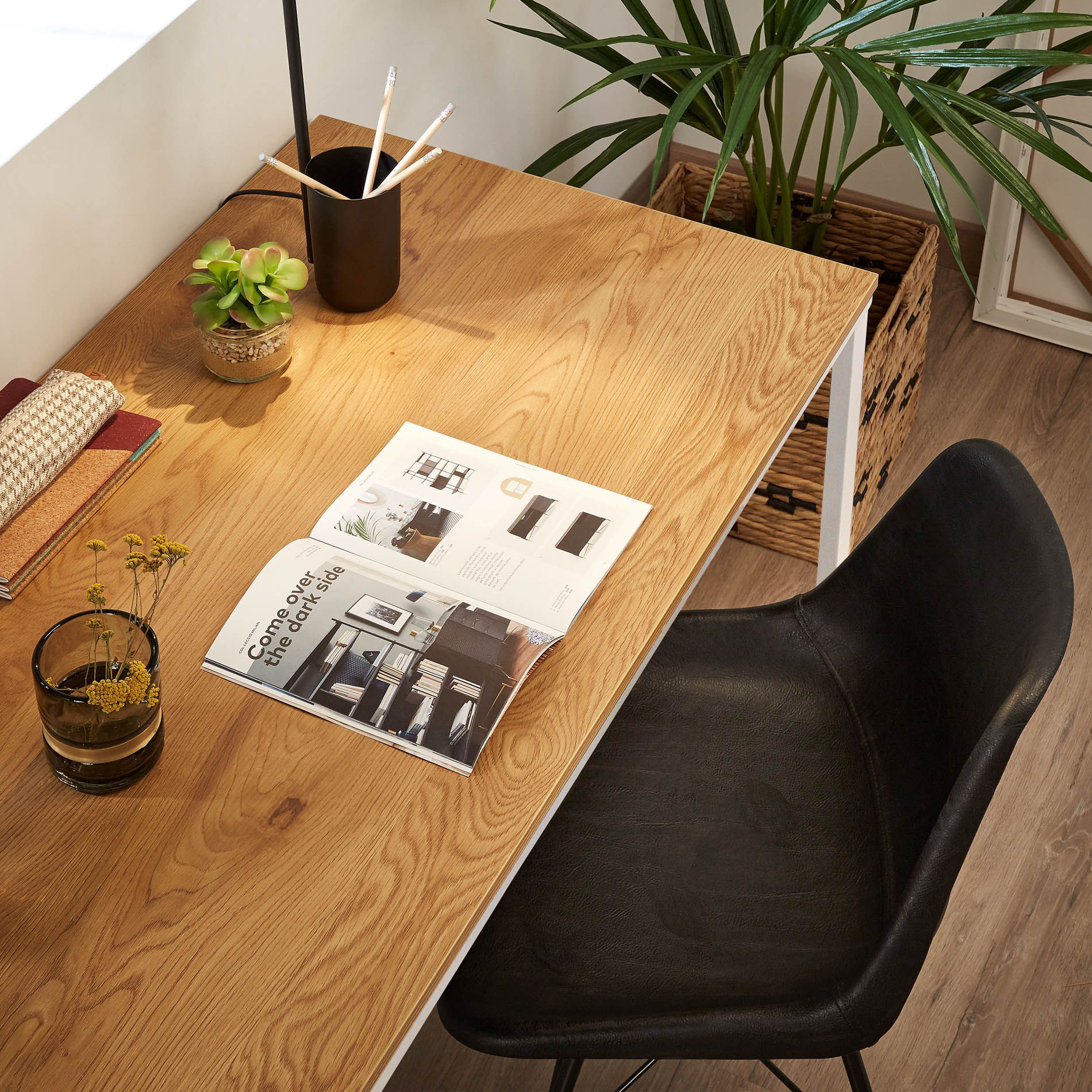 Secretária Talba, MDF/metal, 120x60 Mesa de madeira com cadeira preta, revista aberta, copos e plantas, em ambiente com piso de madeira e planta grande