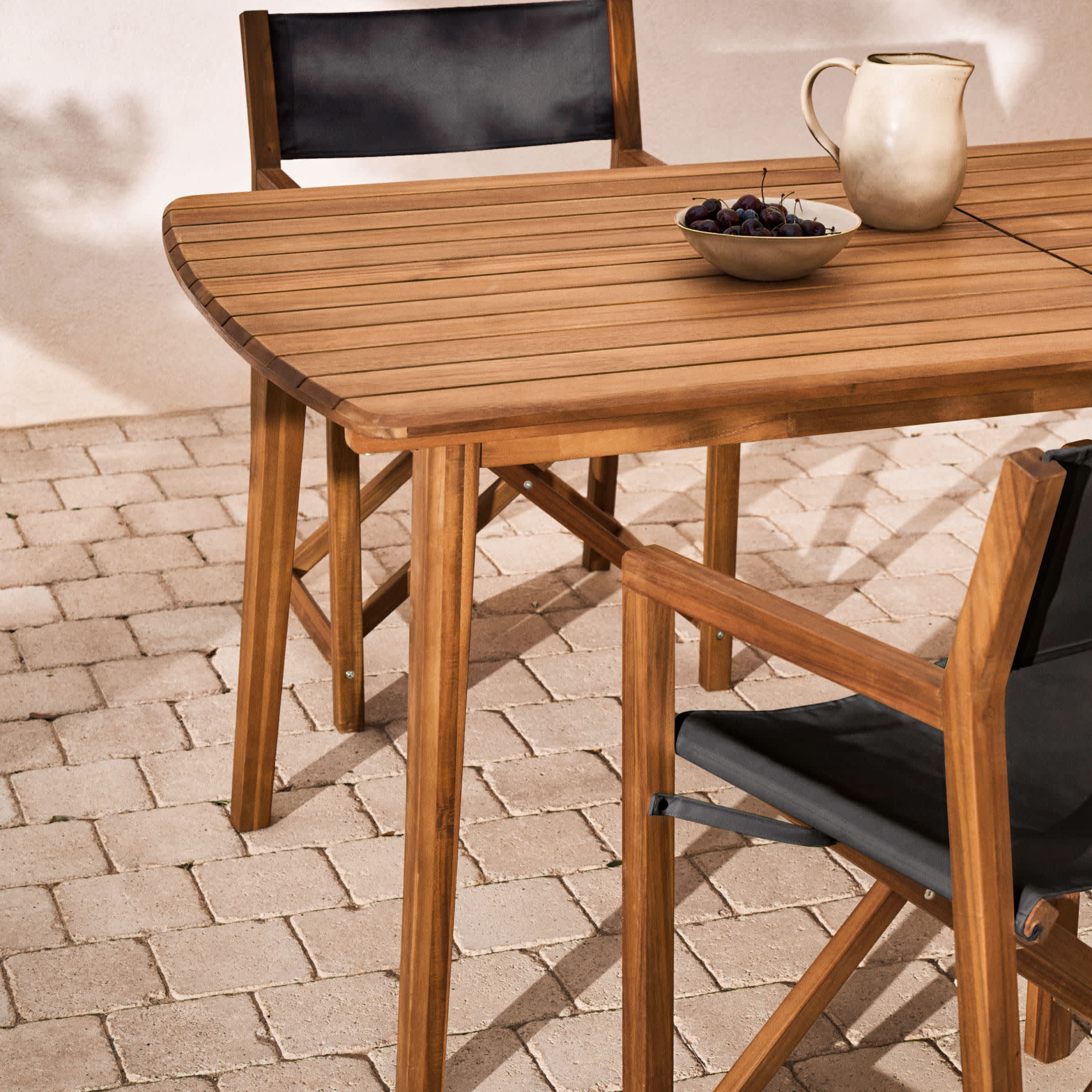 Mesa extensível Thian, exterior, madeira acácia Mesa de madeira clara com cadeiras de tecido preto e jarra branca sobre mesa