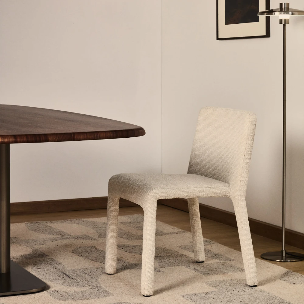 Cadeira empilhável madeira estofada chenille Lorani Cadeira creme tapada, mesa madeira escura, tapete padrão geométrico e candeeiro de pé metálico.