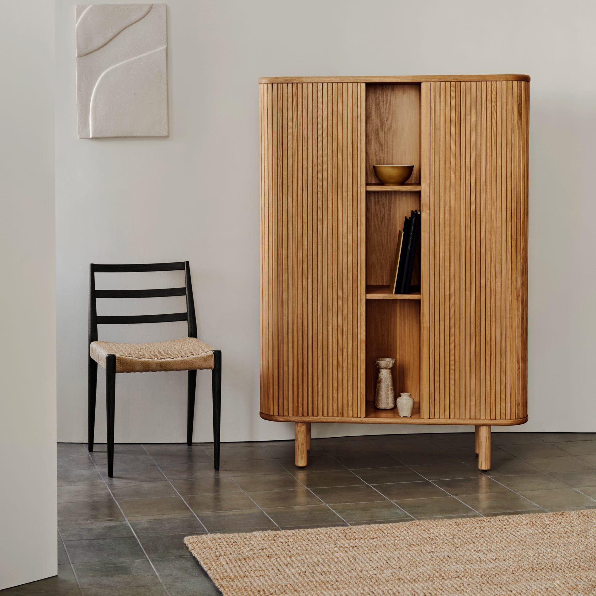 Aparador Malem, madeira de freixo, 110x160 Móvel de madeira com portas ripadas ao lado de cadeira preta com assento de palha em sala com chão cinza