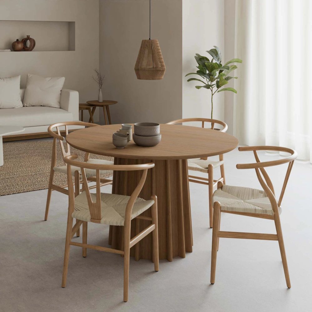 Cadeira com braços em madeira faia Wishbone Mesa de jantar redonda de madeira clara com quatro cadeiras de corda trançada e base de mesa revestida, num ambiente claro com sofá e planta.