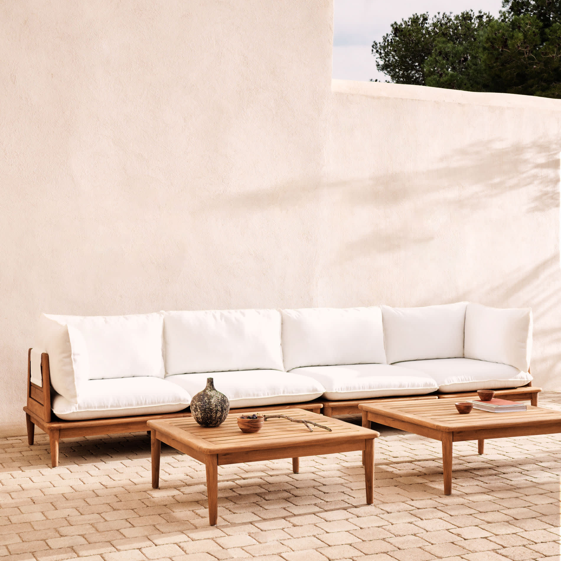 Conjunto Porto Rico, sofá 4 lugares, 2 mesas, madeira teca Sofá de madeira clara com almofadas brancas e mesas de centro em ambiente exterior com parede branca