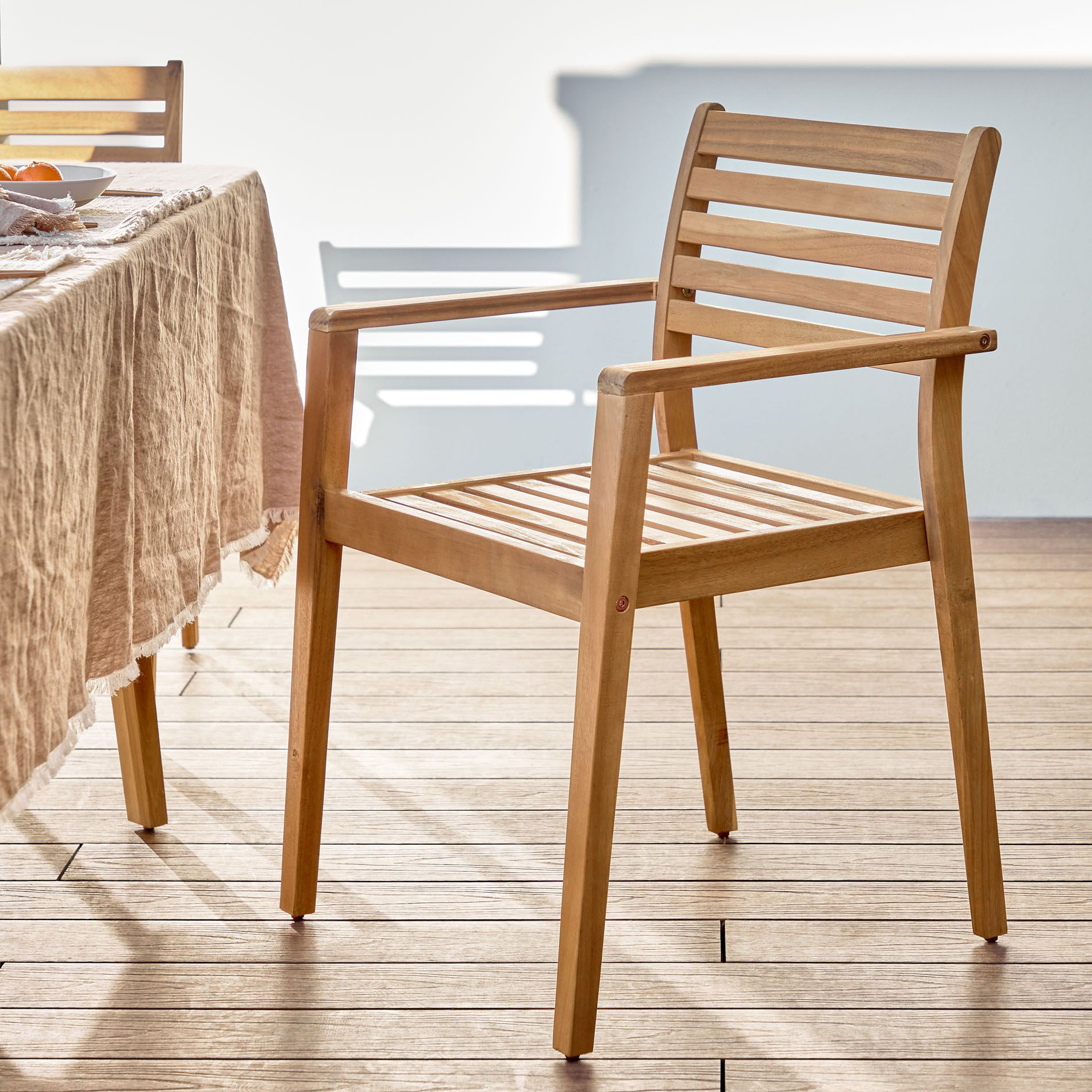 Cadeira Hanz, exterior, madeira acácia Cadeira de madeira clara com ripas horizontais ao lado de mesa com toalha bege
