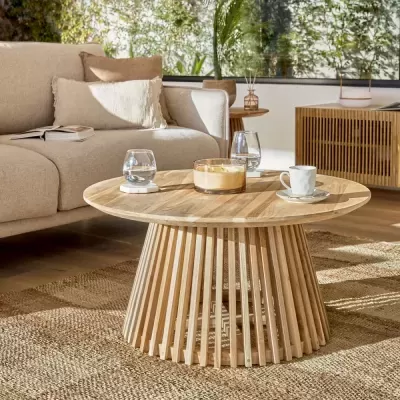 Mesa de centro redonda de madeira clara com copos, vela e chávena sobre tapete em sala com sofá bege