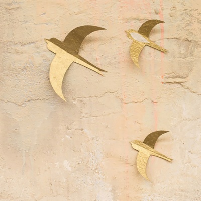 Três figuras douradas de pássaros numa parede bege texturada