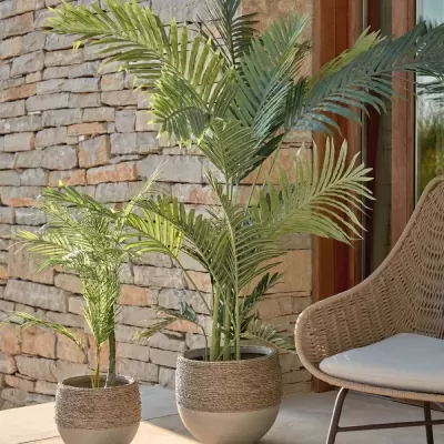 Dois vasos de plantas artificiais com folhas verdes e base texturizada, junto a parede de pedra e cadeira de vime