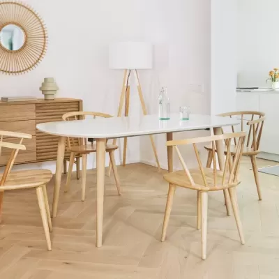 Mesa de jantar branca oval com cadeiras de madeira clara num interior com cómoda e candeeiro de pé